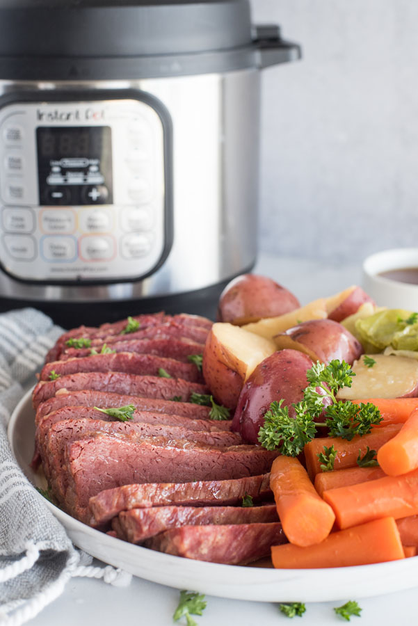 Side view of pressure cooker Corned Beef, cabbage, potatoes and carrots on a white serving platter with an Instant Pot in the background.