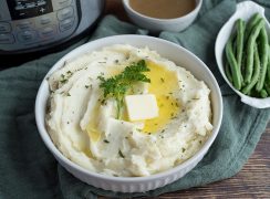 Overhead view of a white bowl of Instapot Mashed Potatoes with a pat of butter on top and an Instant Pot, turkey gravy, and green beans in the background