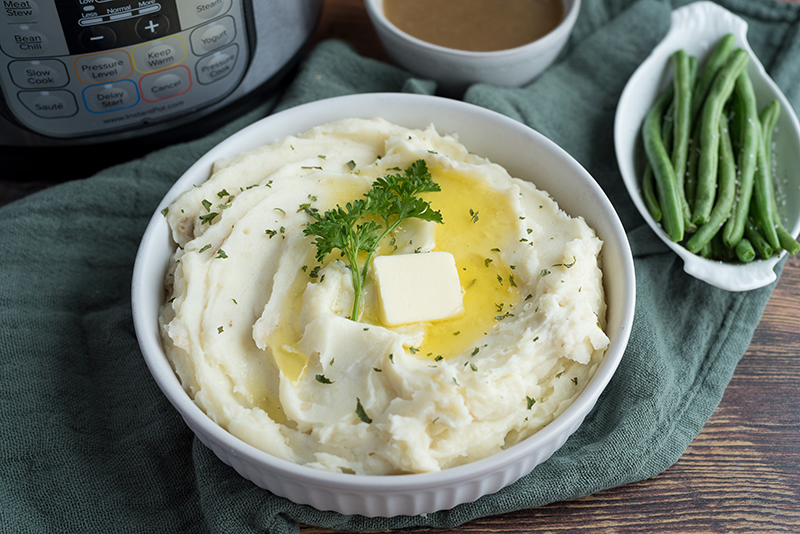 Overhead view of a white bowl of Instapot Mashed Potatoes with a pat of butter on top and an Instant Pot, turkey gravy, and green beans in the background
