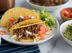 Instant Pot tacos in hard shells on a white plate with tomatoes, onions, cheese, and lettuce, next to bowls with guacamole and tomatoes.
