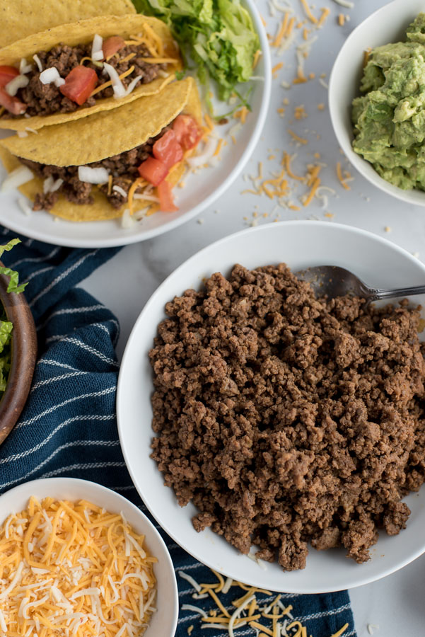 Overhead shot of ground beef taco meat made in and Instant Pot surrounded by a bowl of cheese and guacamole with prepared tacos on a plate.