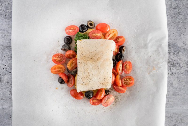 overhead of a fish fillet on a parchment paper with cherry tomatoes and olives under