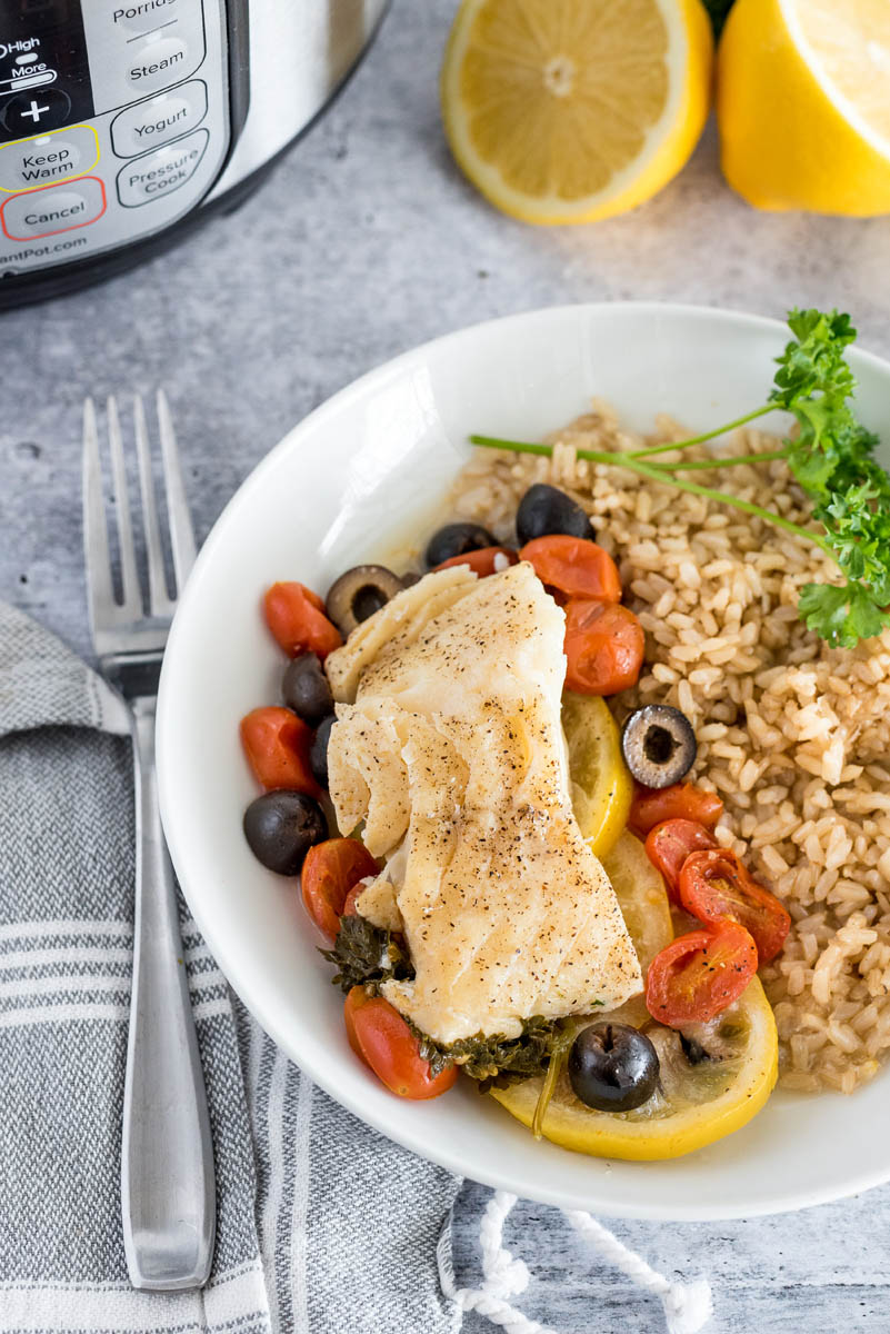 An overhead shot of Instant Pot Fish en Papillote on a white plate with tomatoes, olives and lemons visible underneath, served beside brown rice, with a fork, an Instant Pot, and lemons visible in the background