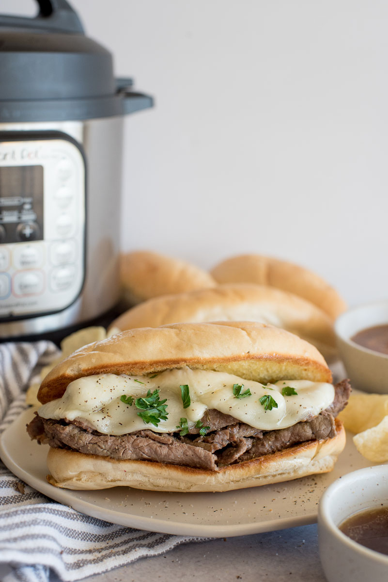 A French dip sandwich topped with cheese on a bun and placed in front of an Instant Pot.