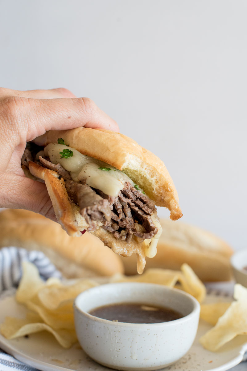 Close up picture of an Instant Pot french dip sandwich topped with cheese and held over a bowl of au jus