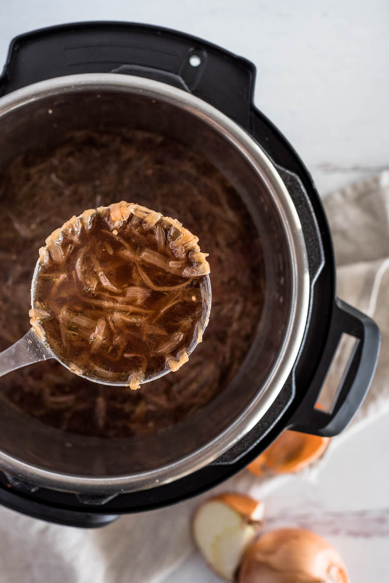 An overhead shot looking into an Instant Pot cooking pot, filled with french onion soup, with a silver ladle taking a spoonful out, with lots of thin onion strips visible