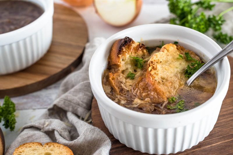 A finished serving of Instant Pot French Onion soup, with browned bread and melted cheese on top of a rich brown french onion soup., served in a white ramekin with a second bowl in the background