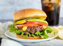 A thick, stacked hamburger, with the patty cooked in the Instant Pot, topped with lettuce, tomato, mustard, pickles, avocado, and a toasted bun on a white plate with chips and a tall glass of cola in the background