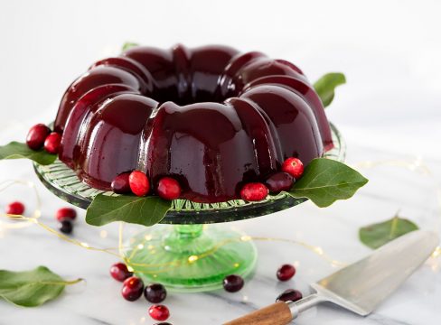 Deep red homemade cranberry jelly in the shape of a bundt pan on top of a green glass cake stand, garnished with bright red cranberries and leaves