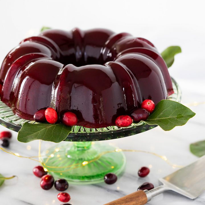 Deep red homemade cranberry jelly in the shape of a bundt pan on top of a green glass cake stand, garnished with bright red cranberries and leaves