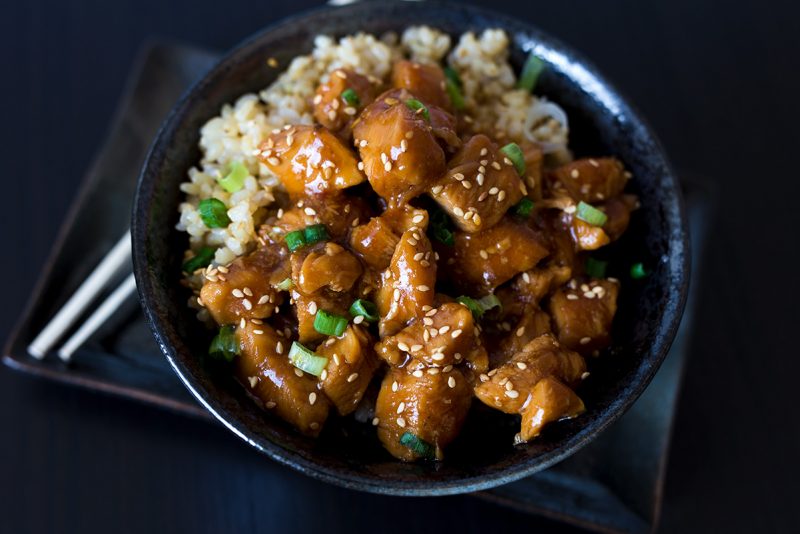 A horizontal close-up of Honey Sesame Chicken in a dark black bowl against a dark black background, with chopsticks tucked under the bowl