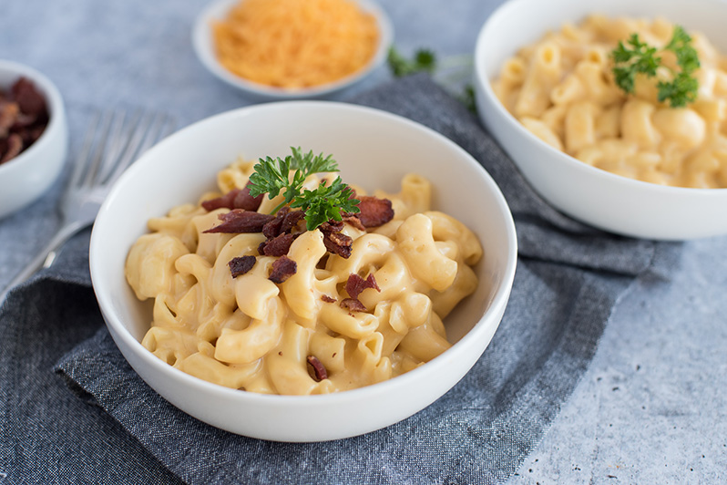 Two bowls of homemade pressure cooker macaroni and cheese topped with fresh parsley and crumbly bacon with a bowl of shredded cheddar cheese in the background.