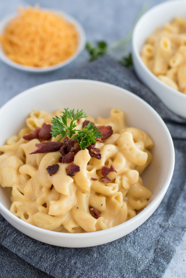 Close up on pressure cooker macaroni and cheese in a white bowl topped with crumbled bacon and fresh parsley with a bowl of shredded cheddar cheese in the background.