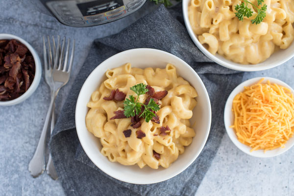 Over head of a bowl of creamy Instant Pot mac and cheese topped with crumbled bacon and fresh parsley with a bowl of shredded cheddar cheese and more bacon for topping on a grey background.