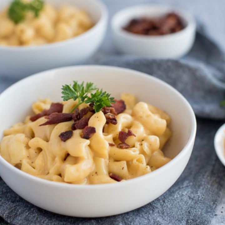 Close up on a white bowl of homemade macaroni and cheese topped with bacon and fresh parsley next to a bowl of shredded cheddar cheese.