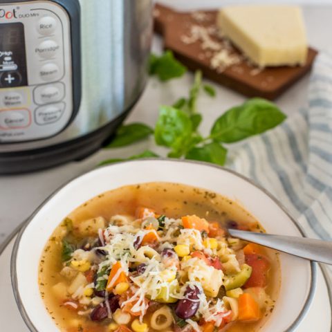 45 degree angle of pressure cooker minestrone soup in a gold rimmed white bowl, with basil, parmesan, and an Instant Pot in the background.