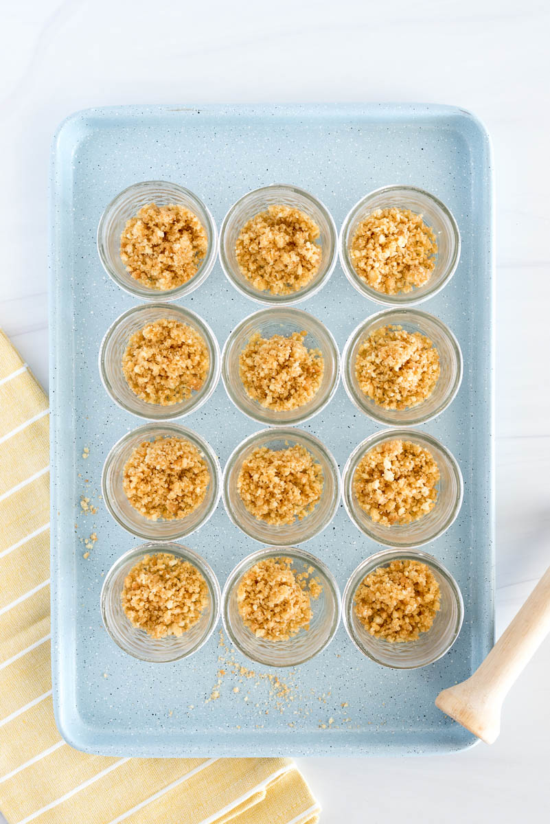 An overhead shot of twelve mini mason jars sitting on top of a blue cookie sheet, filled with crushed coconut cookies, with a bright yellow napkin and a tart tamper in the background