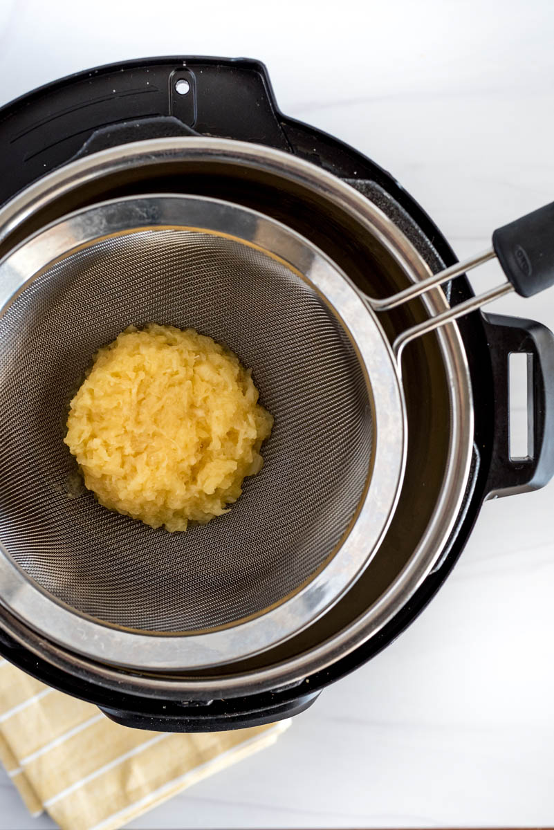 An overhead shot of crushed pineapple draining through a fine mesh sieve into the cooking pot of an Instant Pot