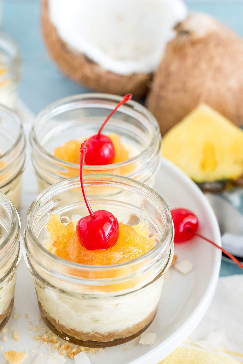 Close-up of Instant Pot Coconut Cheesecakes with a pineapple topping, sitting on a white plate, with a coconut and pineapple wedge in the background
