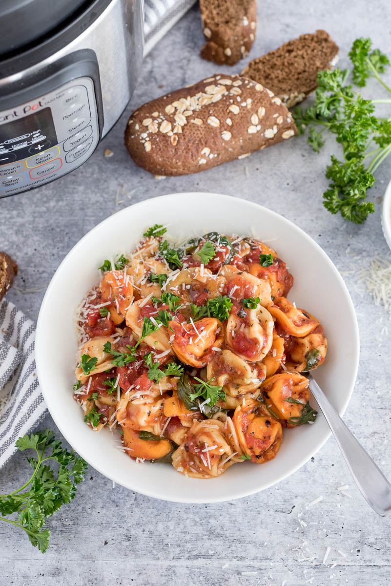 Easy tortellini cooked in a tomato basil sauce topped with fresh parsley and parmesan and placed in a white bowl in front of an Instant Pot.