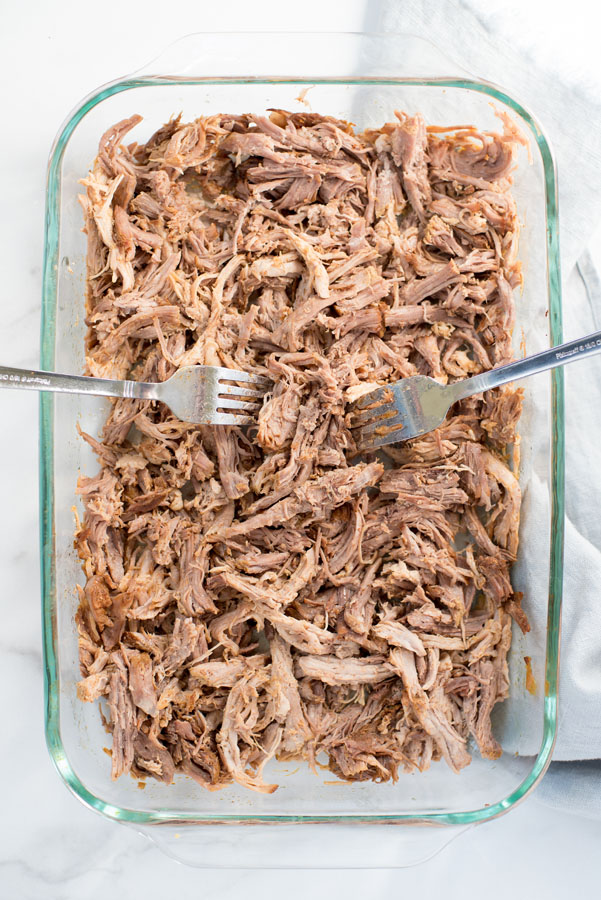 Overhead of a glass baking dish filled with pressure cooked pork shoulder being shredded with two silver forks to make filling for homemade pork tamales.