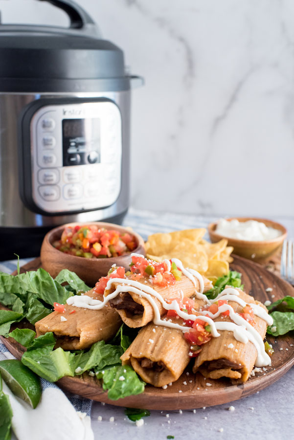 Wooden serving plate of four Instant Pot pork tamales made with masa and wrapped in corn husks with sour cream, fresh salsa, lettuce, tortilla chips and limes with an electric pressure cooker in the background.