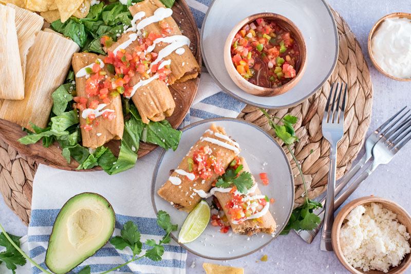 Overhead shot of Instant Pot Pork Tamales, featuring a large serving platter with tamales, two tamales plated up, and a bowl full of servings, with cilantro and avocado on the page
