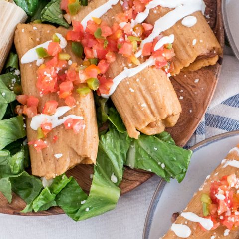 Overhead close up of several pork tamales, laying on a bed of lettuce, topped with tomatoes and sour cream, with an avocado in the bottom left corner