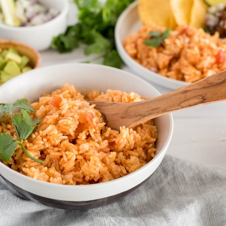 45 degree shot of Instant Pot spanish rice with a wooden serving spoon, garnished with parsley with a background of boowls of avocado, onions, and plate with beef tacos in the background
