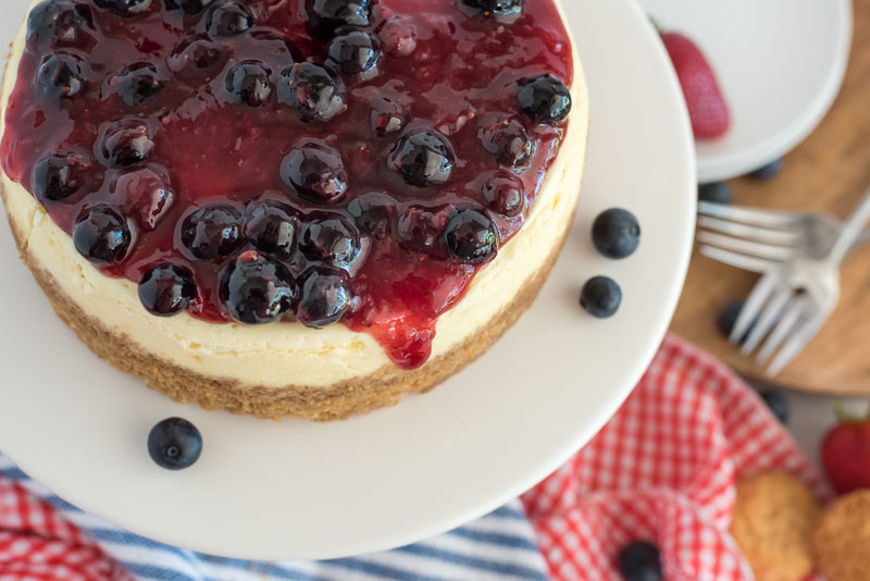 Close up overhead shot of Instant Pot cheesecake topped with berry topping sitting on a white cake stand with red, white, and blue napkins in the background.