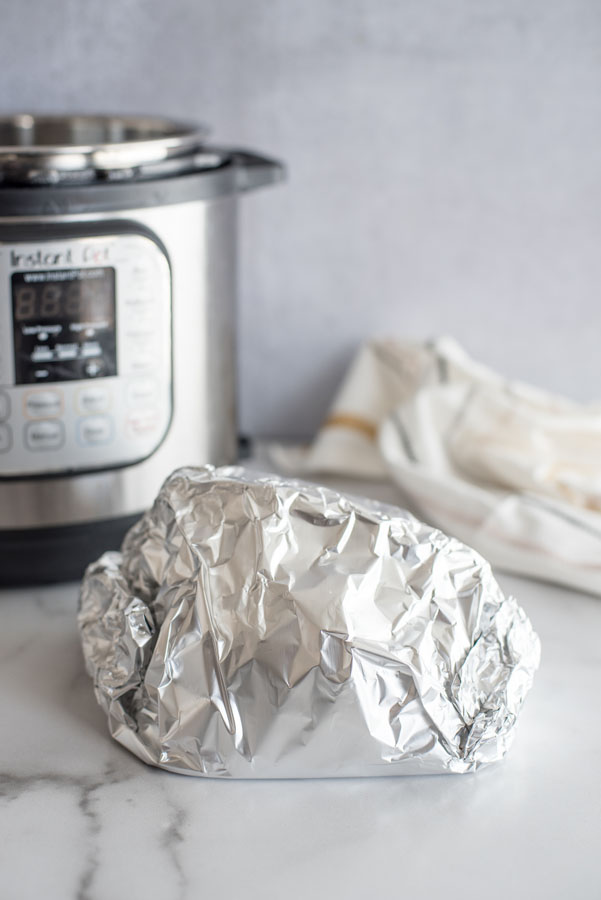 Ham wrapped in aluminum foil to be heated in the Instant Pot / pressure cooker on a marble counter with a white cloth in the background.