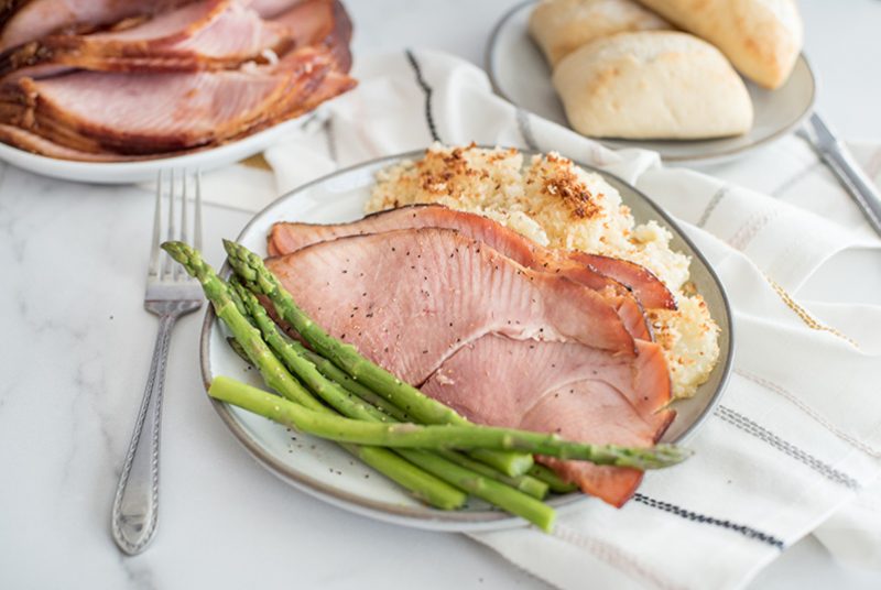 45 degree overhead of warmed spiral ham slices served with crisp green asparagus and mashed potatoes made in the pressure cooker on a white plate with fresh rolls in the background.