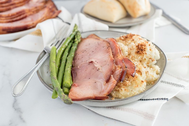 Overhead of a white dish with crisped potato gratin, two slices of pink Easter ham, and fresh asparagus spears with more ham and fresh dinner rolls in the background.