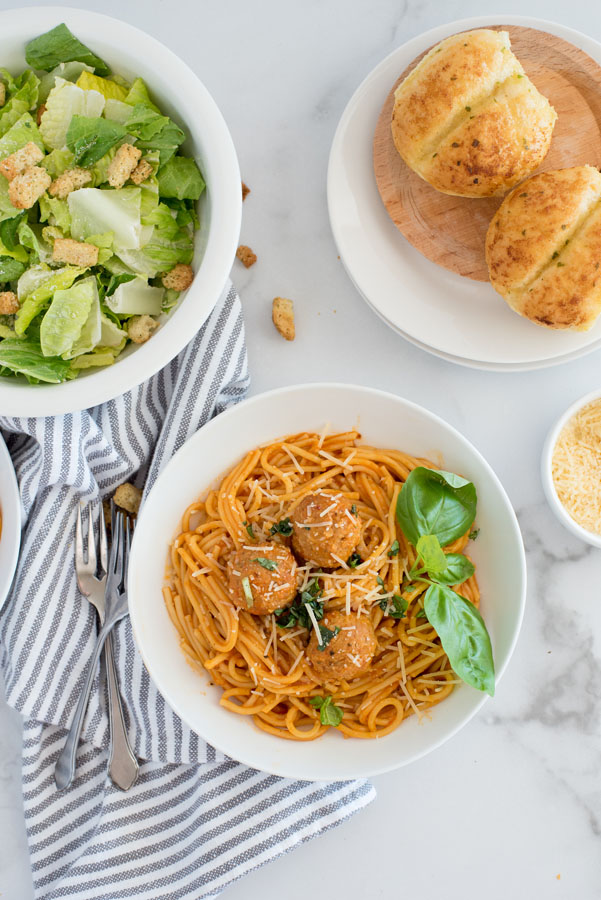 flat lay of a white pasta dish filled with isntant pot spaghetti and meatballs served with a caesar salad and fresh bread rolls on a white marble background