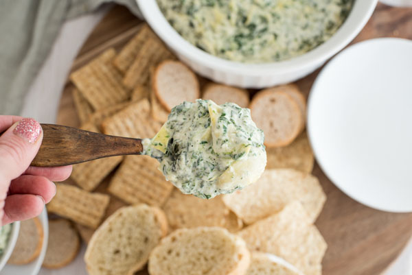 Smooth and creamy spinach artichoke dip being scooped into a bowl next to a platter of crackers.