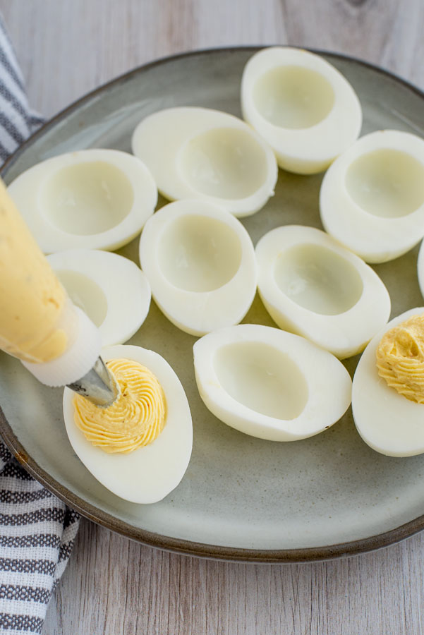 Close up on a dozen hard-boiled egg whites sliced in half on a ceramic plate being filled with deviled egg filling from a pastry bag with a star tip.
