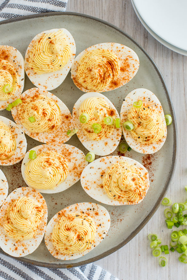 Overhead of a ceramic plate filled with Instant Pot deviled eggs sprinkled with smoked paprika on a light wooden background with sliced scallions on the side.