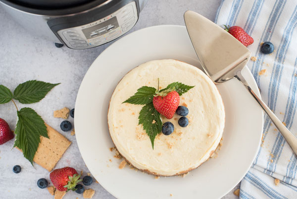 A top view of Instant Pot cheesecake, garnished with a strawberry and blueberries and mint leaves, with a silver server and an Instant Pot in the background