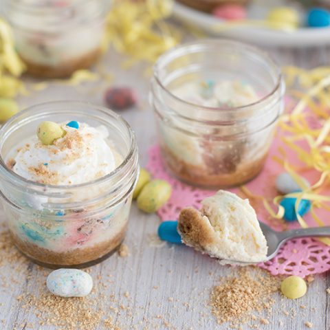 Side view of two mini pressure cooker Robin Egg’s Cheesecake made in mini glass mason jars with Whoppers Robin Eggs on a white wooden table decorated for Easter.