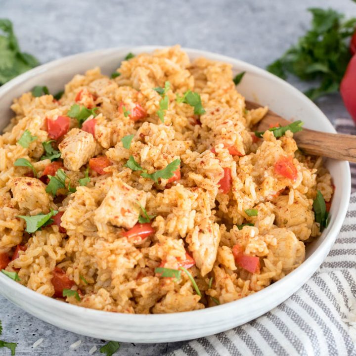 A 45 degree shot of a white bowl filled with Spanish style Instant Pot chicken and rice, garnished with red bell peppers and diced parsley, with a wooden spoon tucked in the back right of the bowl, with extra parsley and red bell peppers in the background