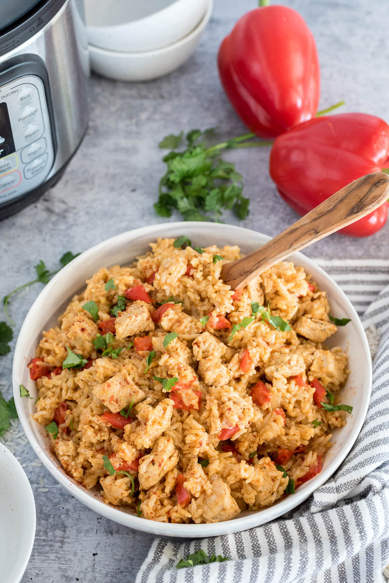 45 degree shot of a white bowl full of Instant Pot spanish style chicken and rice, with an instant pot, bell peppers, and cilantro in the background
