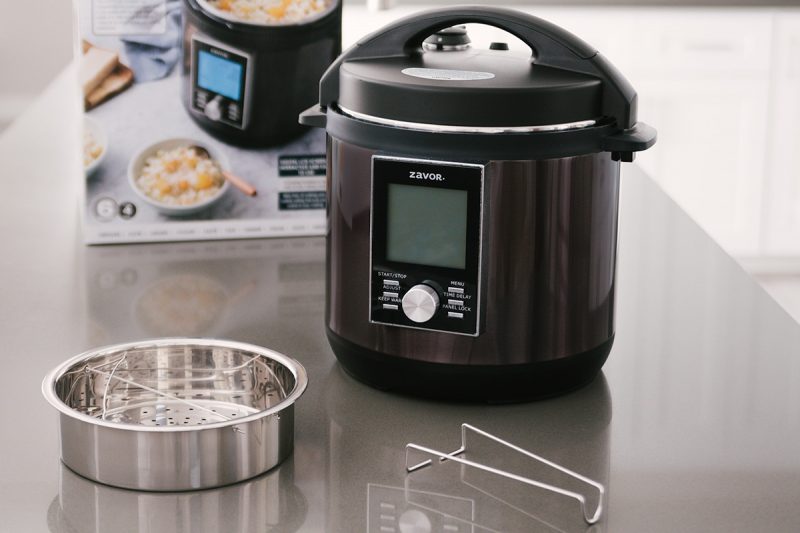 The Zavor LUX pressure cooker placed on a counter with the included trivet and steamer basket placed in front of the packaging box.