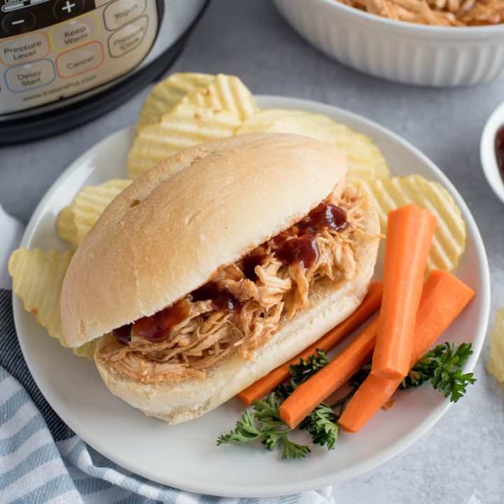 A 45 degree shot of a BBQ chicken sandwich, with carrots and potato chips in front of an Instant Pot with additional shredded chicken in a white bowl in the background
