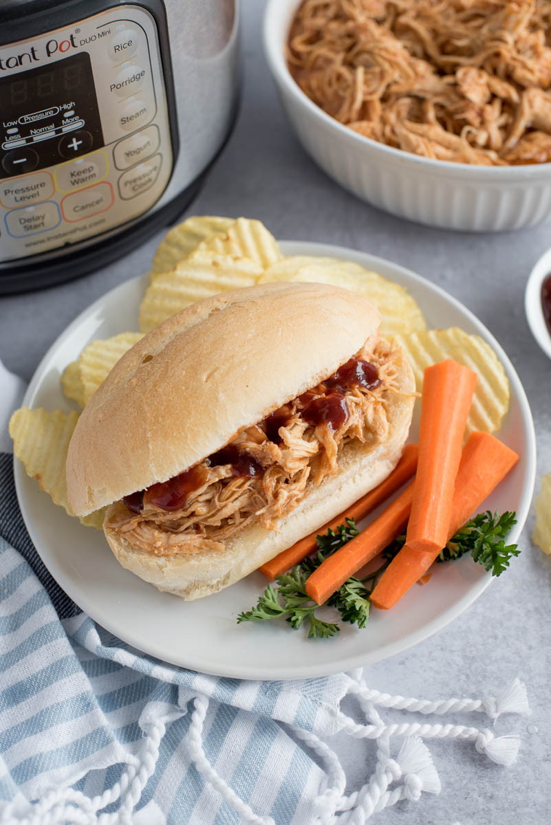 A 45 degree shot of a BBQ chicken sandwich, with carrots and potato chips in front of an Instant Pot with additional shredded chicken in a white bowl in the background