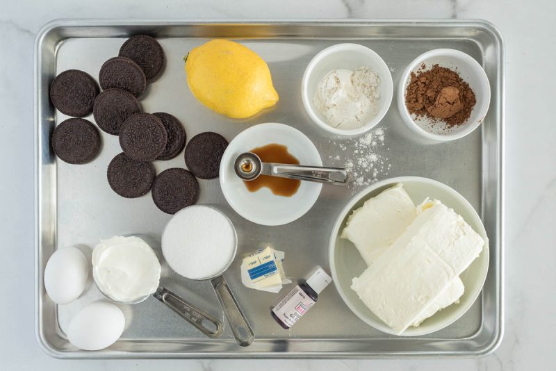 an overhead shot of the ingredients needed to make red velvet cheesecake, including Oreo cookies, lemon juice, flour, cocoa, vanilla, cream cheese, sour cream, eggs, butter, sugar, and red food coloring