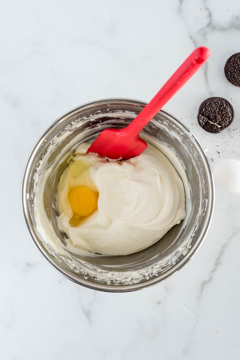 an overhead shot of a silver mixing bowl and a red spatula showing the cream cheese and sugar until just combined, then adding the eggs one at a time