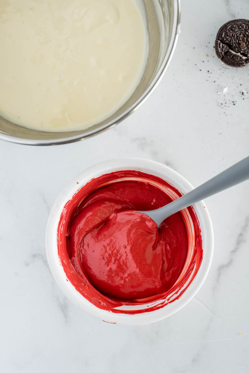 An overhead shot looking into two bowls. A smaller white bowl, showing the red food coloring mixed in with a gray spatula, and larger silver bowl filled with the remaining white cheesecake batter.