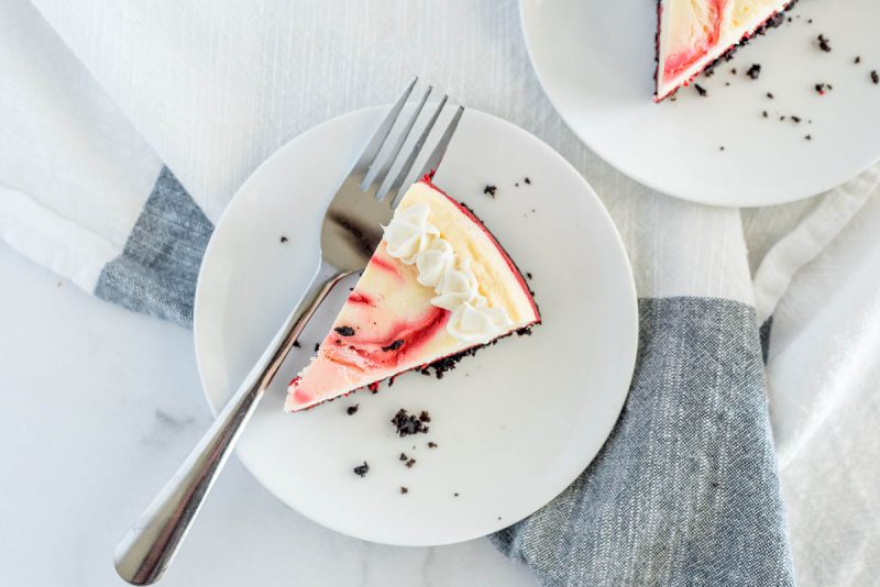 A slice of red velvet cheesecake on a small white plate with a fork, garnished with chocolate crumbs and finished with a whipped cream border