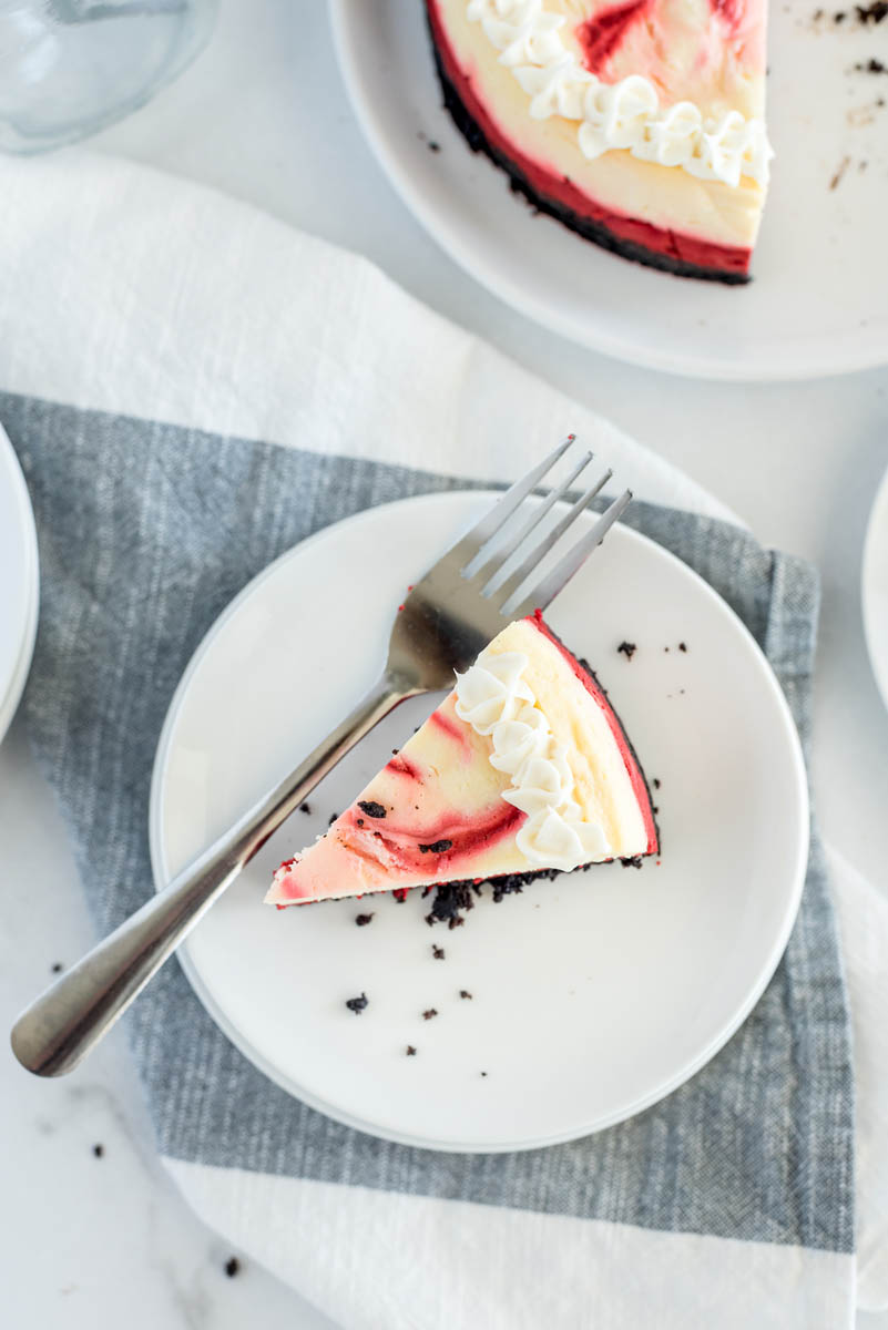 An overhead shot of Instant Pot red velvet cheesecake, sitting on a white plate and a gray linen napkin, with the rest of the cheesecake visible on the top right