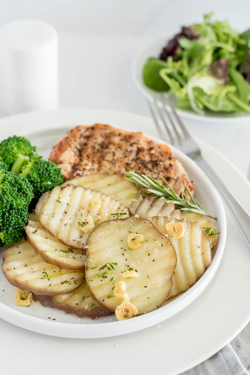 a 45 degree shot of a plate with new potatoes drizzled with sliced garlic, rosemary, salt, on a white plate with broccoli and chicken with a small side salad visible in the background.